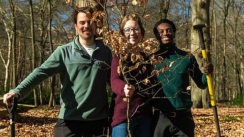 Drie personen staan in een bos met een schep en een uitgegraven jong boompje in hun handen. Alle drie hebben ze een glimlach op hun gezicht. 